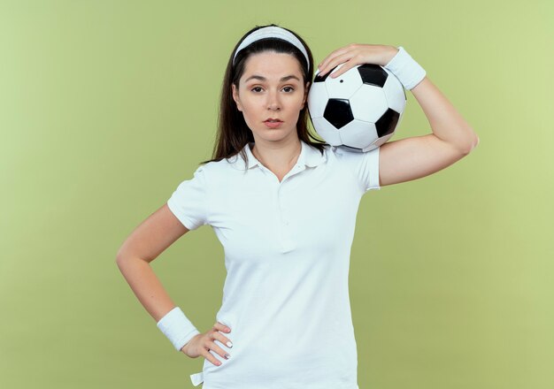 Mujer joven fitness en diadema sosteniendo un balón de fútbol en su hombro mirando a la cámara con expresión de confianza de pie sobre fondo claro