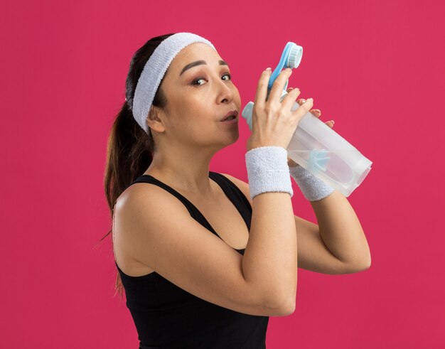 Mujer joven fitness con diadema sosteniendo una botella de agua con cara seria