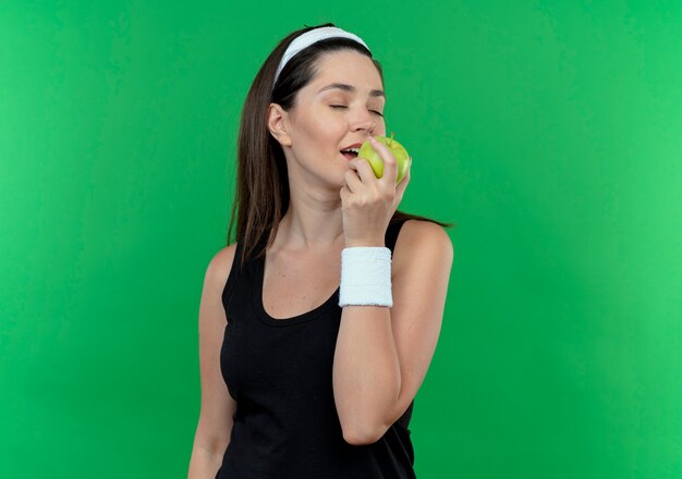 Mujer joven fitness en diadema sosteniendo manzana verde mordiendo feliz y positivo de pie sobre fondo verde