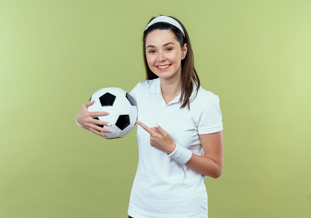 Mujer joven fitness en diadema sosteniendo una pelota de fútbol apuntando con el dedo sonriendo alegremente de pie sobre la pared de luz