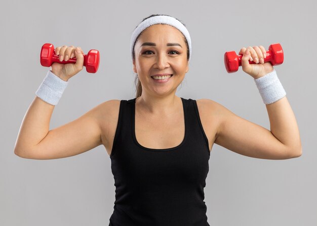 Mujer joven fitness con diadema sosteniendo pesas haciendo ejercicios mirando seguro sonriendo de pie sobre la pared blanca