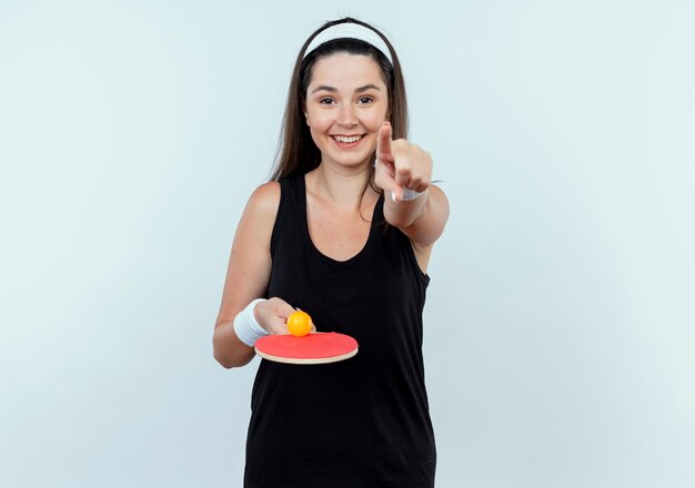 Mujer joven fitness en diadema sosteniendo la raqueta y la pelota de tenis de mesa apuntando con el dedo sonriendo alegremente de pie sobre la pared blanca