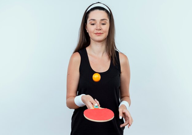 Mujer joven fitness en diadema sosteniendo la raqueta y la pelota de tenis de mesa lanzando una pelota sonriendo confiados de pie sobre la pared blanca