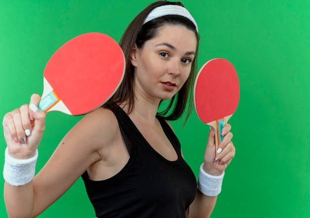 Mujer joven fitness en diadema sosteniendo raquetas de tenis de mesa mirando a la cámara con expresión de confianza de pie sobre fondo verde