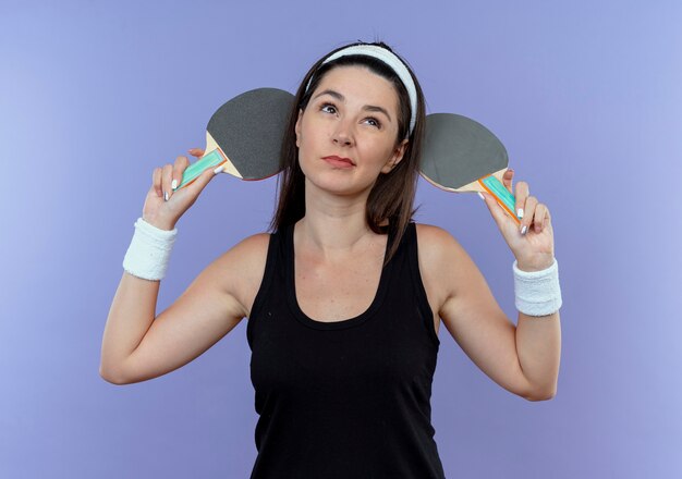 Mujer joven fitness en diadema sosteniendo raquetas de tenis de mesa mirando desconcertado parado sobre fondo azul.