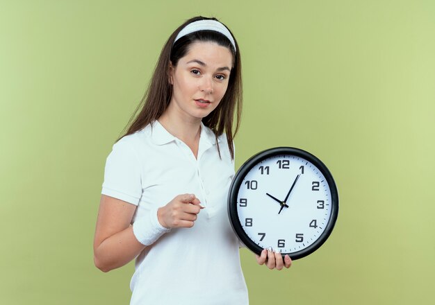 Mujer joven fitness en diadema sosteniendo reloj de pared apuntando con el dedo sonriendo de pie sobre la pared de luz