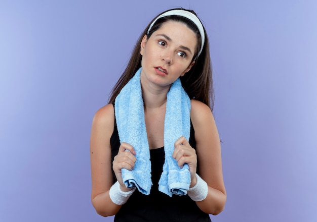 Mujer joven fitness en diadema con una toalla alrededor del cuello mirando a un lado cansado y aburrido de pie sobre fondo azul.
