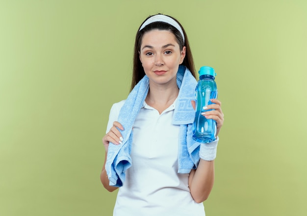 Foto gratuita mujer joven fitness en diadema con una toalla alrededor del cuello sosteniendo una botella de agua mirando a la cámara sonriendo confiados de pie sobre fondo claro