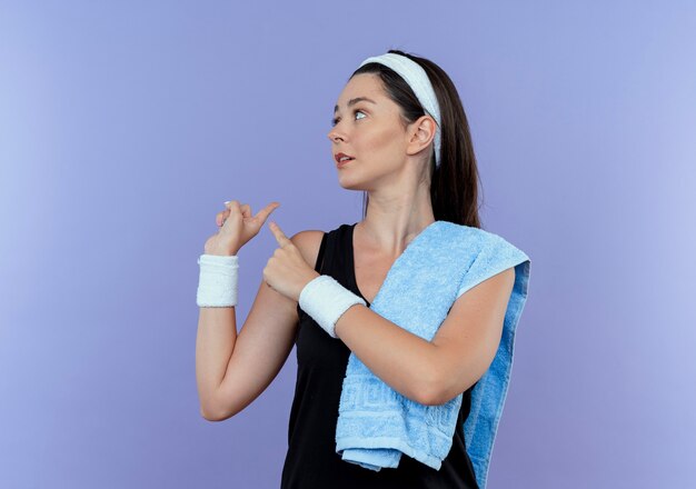 Mujer joven fitness en diadema con una toalla en el hombro mirando a un lado apuntando hacia atrás de pie sobre la pared azul