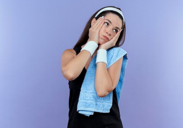 Foto gratuita mujer joven fitness en diadema con una toalla en el hombro mirando a un lado molesto soplar las mejillas de pie sobre fondo azul.