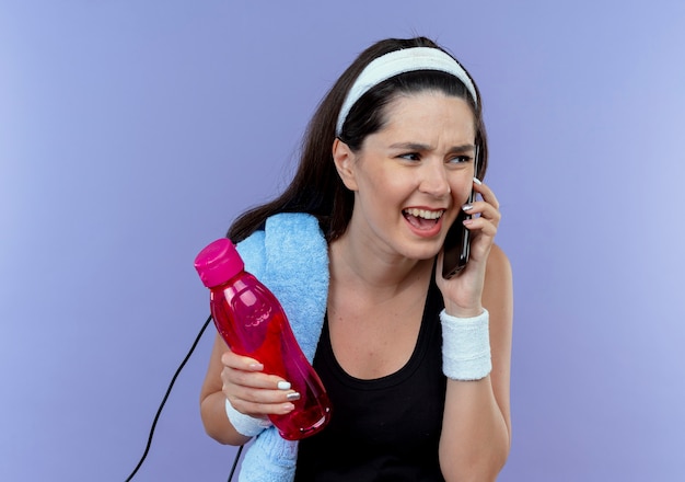 Mujer joven fitness en diadema con una toalla en el hombro sosteniendo una botella de agua hablando por teléfono móvil con expresión molesta de pie sobre fondo azul