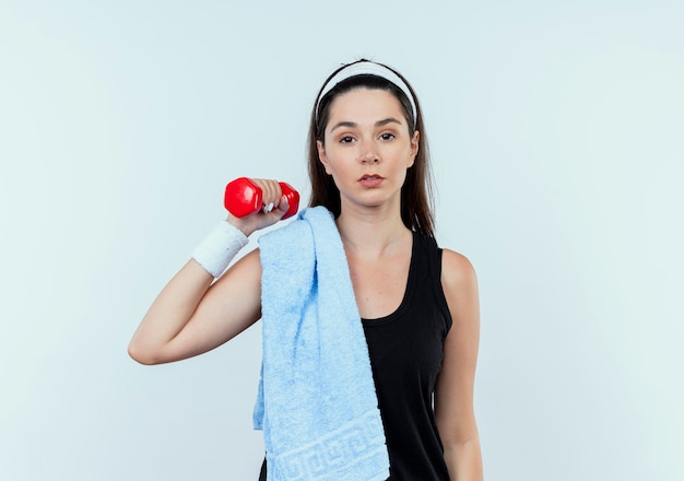 Mujer joven fitness en diadema con una toalla sobre su hombro trabajando con mancuernas mirando confiado de pie sobre fondo blanco.