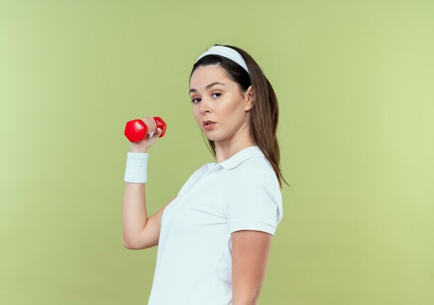 Mujer joven fitness en diadema trabajando con mancuernas con cara seria de pie sobre una pared de luz