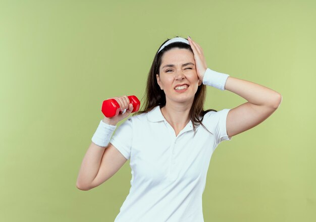 Mujer joven fitness en diadema trabajando con mancuernas mirando confundido parado sobre pared de luz