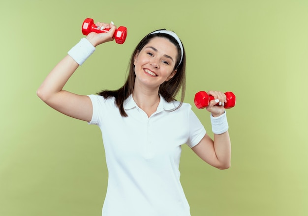 Mujer joven fitness en diadema trabajando con pesas mirando confiado sonriendo feliz y positivo de pie sobre fondo claro