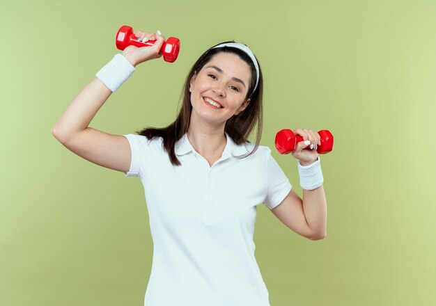 Mujer joven fitness en diadema trabajando con pesas mirando confiado sonriendo feliz y positivo de pie sobre la pared de luz