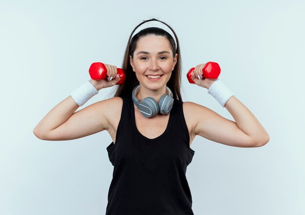 Mujer joven fitness en diadema trabajando con pesas mirando confiado sonriendo de pie sobre fondo blanco.