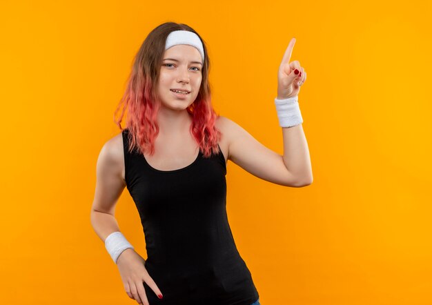 Mujer joven fitness en ropa deportiva apuntando con el dedo hacia el lado parado sobre la pared naranja