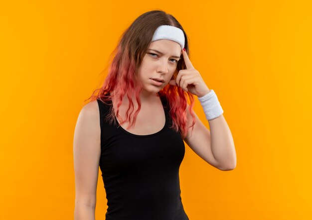 Mujer joven fitness en ropa deportiva apuntando su templo concentrándose en una tarea mirando confiado parado sobre la pared naranja