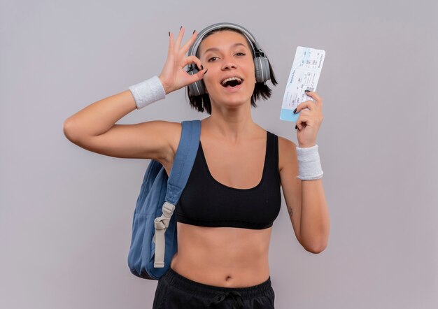 Mujer joven fitness en ropa deportiva con auriculares en la cabeza con mochila sosteniendo boleto aéreo haciendo bien firmar sonriendo alegremente de pie sobre la pared blanca