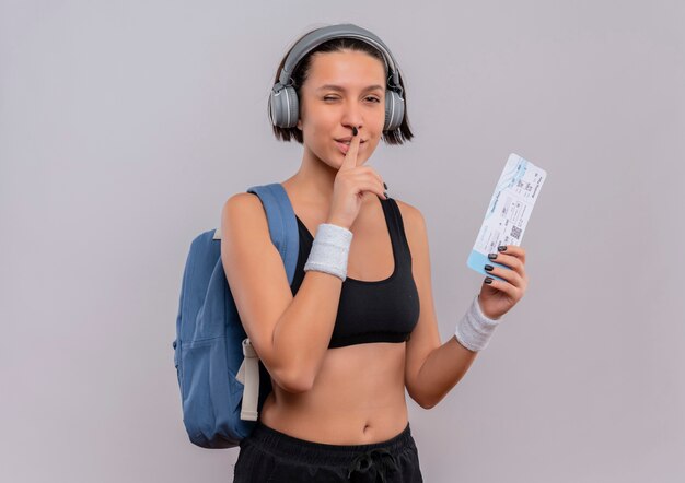 Mujer joven fitness en ropa deportiva con auriculares en la cabeza con mochila sosteniendo boleto aéreo haciendo gesto de silencio con el dedo en los labios