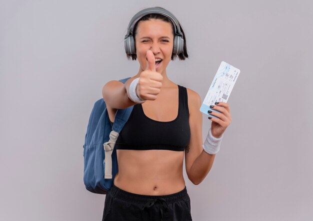 Mujer joven fitness en ropa deportiva con auriculares en la cabeza con mochila sosteniendo boleto aéreo sonriendo mostrando los pulgares para arriba de pie sobre la pared blanca