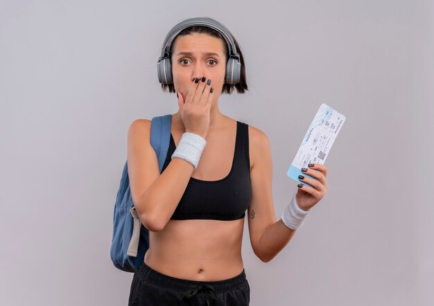 Mujer joven fitness en ropa deportiva con auriculares en la cabeza con mochila sosteniendo boleto aéreo sorprendido cubriendo la boca con la mano de pie sobre la pared blanca