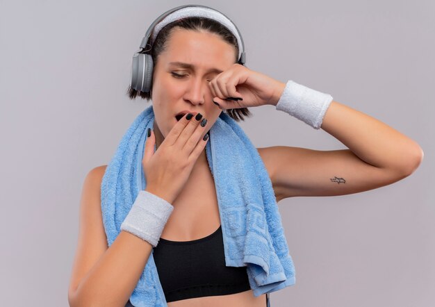 Mujer joven fitness en ropa deportiva con auriculares en la cabeza y una toalla en el cuello bostezando cubriendo la boca con la mano quiere dormir de pie sobre la pared blanca