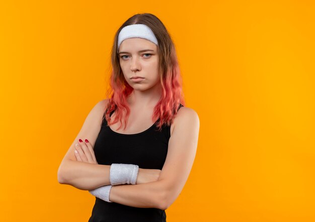 Mujer joven fitness en ropa deportiva con los brazos cruzados sobre el pecho con cara seria de pie sobre la pared naranja