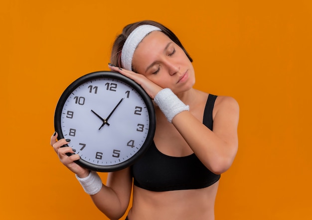 Mujer joven fitness en ropa deportiva con diadema abrazando reloj de pared con la cabeza inclinada sobre él con los ojos cerrados de pie sobre la pared naranja