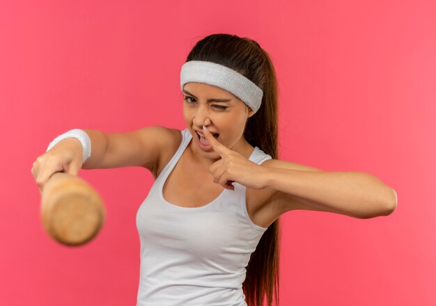 Mujer joven fitness en ropa deportiva con diadema apuntando con un bate de béisbol guiñando un ojo y sonriendo de pie sobre la pared rosa