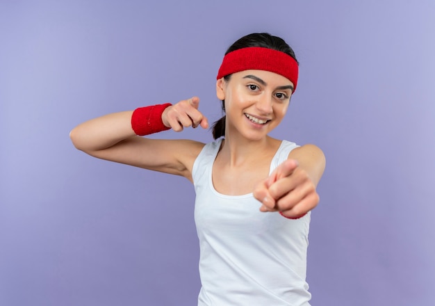 Mujer joven fitness en ropa deportiva con diadema apuntando con los dedos índices a la cámara sonriendo alegremente de pie sobre la pared púrpura