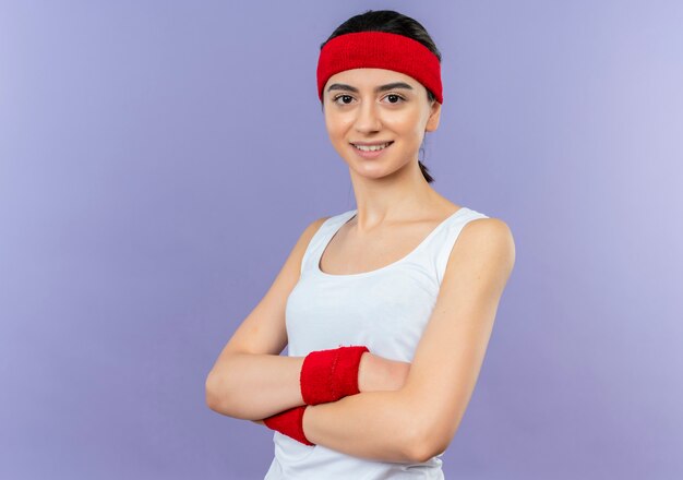 Mujer joven fitness en ropa deportiva con diadema con los brazos cruzados sobre el pecho sonriendo confiado de pie sobre la pared púrpura