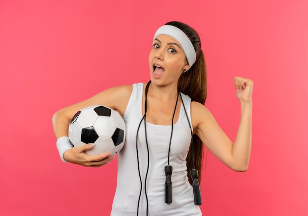Mujer joven fitness en ropa deportiva con diadema y cuerda alrededor de su cuello sosteniendo un balón de fútbol mirando sorprendido