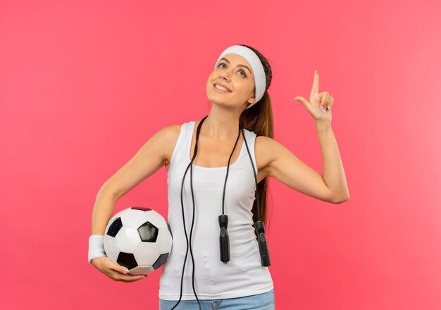 Mujer joven fitness en ropa deportiva con diadema y cuerda alrededor de su cuello sosteniendo un balón de fútbol