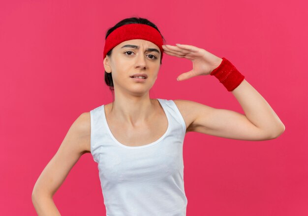 Mujer joven fitness en ropa deportiva con diadema con expresión escéptica saludando de pie sobre la pared rosa