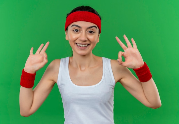 Mujer joven fitness en ropa deportiva con diadema feliz y positiva sonriendo alegremente haciendo bien firmar de pie sobre la pared verde