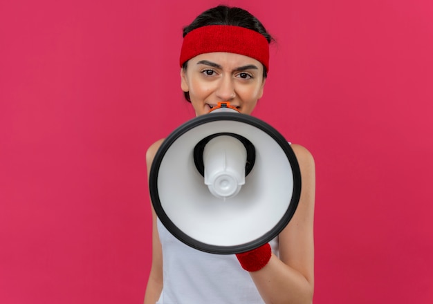 Mujer joven fitness en ropa deportiva con diadema gritando al megáfono con cara enojada de pie sobre la pared rosa