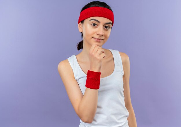 Foto gratuita mujer joven fitness en ropa deportiva con diadema con la mano en la barbilla mirando confiado de pie sobre la pared púrpura