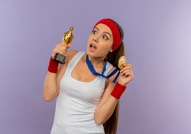 Mujer joven fitness en ropa deportiva con diadema con medalla de oro alrededor de su cuello sosteniendo su trofeo mirando sorprendido