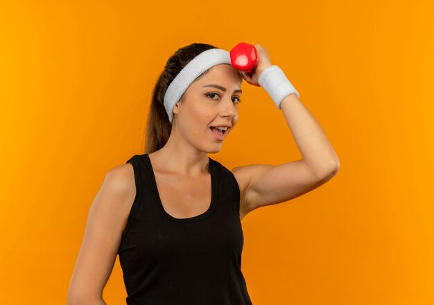 Mujer joven fitness en ropa deportiva con diadema mirando confiado sosteniendo mancuernas de pie sobre pared naranja