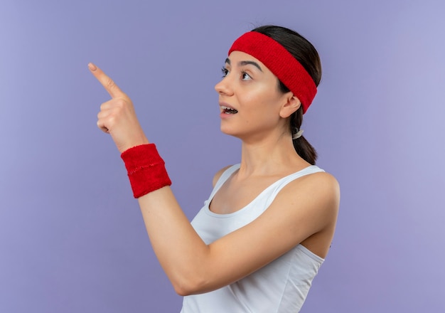 Foto gratuita mujer joven fitness en ropa deportiva con diadema mirando a un lado sorprendido y asombrado señalando con el dedo hacia el lado de pie sobre la pared púrpura