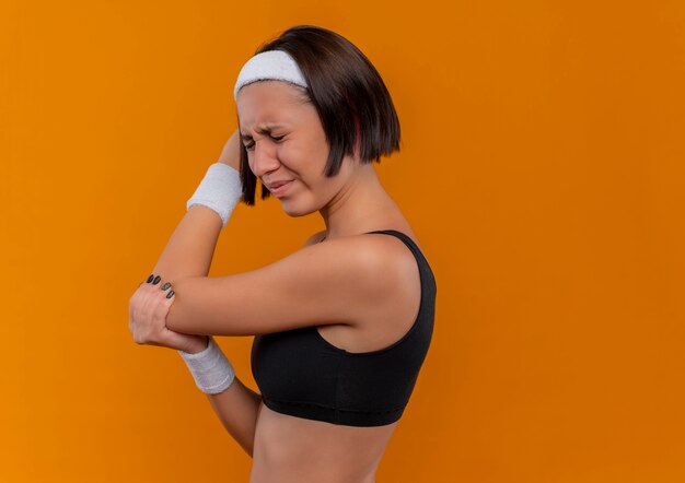 Mujer joven fitness en ropa deportiva con diadema mirando mal tocando su codo sintiendo dolor de pie sobre la pared naranja