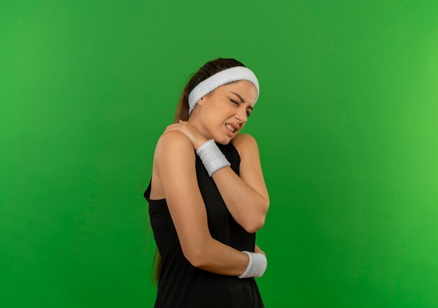 Mujer joven fitness en ropa deportiva con diadema mirando mal tocando su hombro con dolor de pie sobre la pared verde