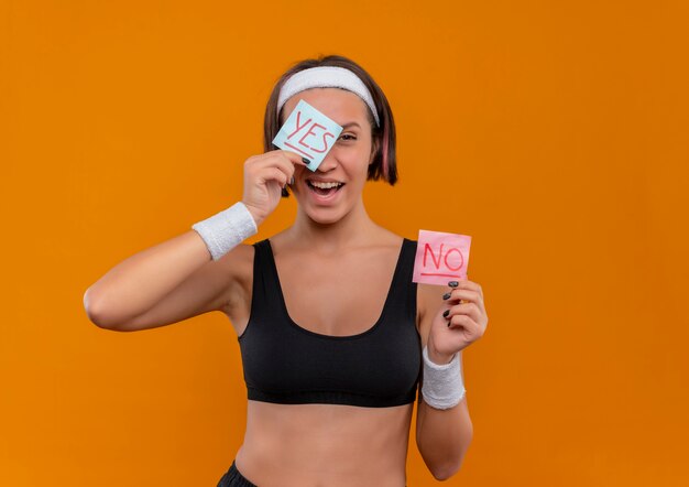 Mujer joven fitness en ropa deportiva con diadema mostrando dos papeles recordatorios con la palabra sí y no sonriendo alegremente cubriendo con papel un ojo parado sobre una pared naranja