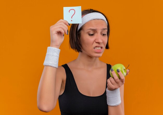 Mujer joven fitness en ropa deportiva con diadema mostrando papel recordatorio con signo de interrogación sosteniendo manzana verde mirándola confundida y muy ansiosa de pie sobre la pared naranja