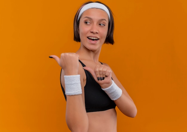 Mujer joven fitness en ropa deportiva con diadema positiva y feliz apuntando hacia atrás con los pulgares de pie sobre la pared naranja