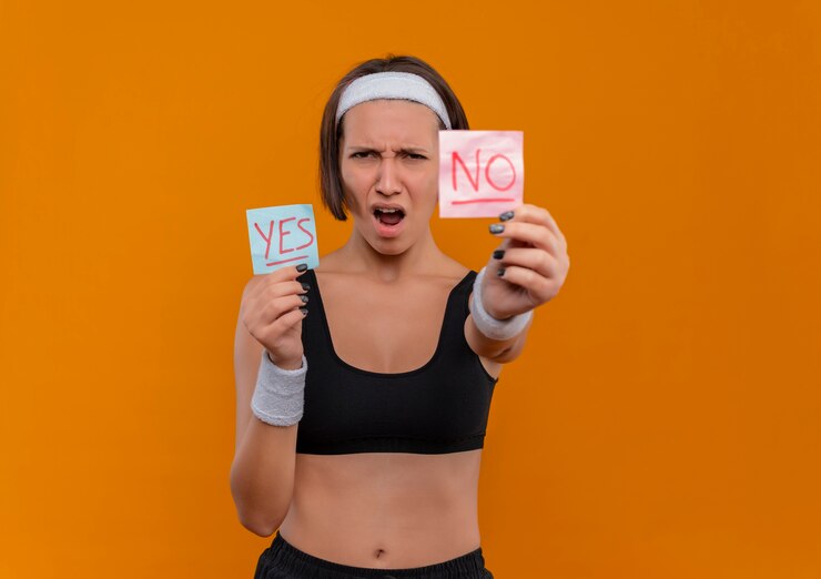 Mujer joven fitness en ropa deportiva con diadema que muestra dos papeles recordatorios con la palabra sí y no sonriendo alegremente de pie sobre la pared naranja