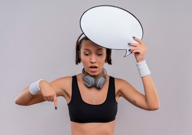 Mujer joven fitness en ropa deportiva con diadema con signo de burbuja de discurso en blanco apuntando con el dedo hacia abajo mirando sorprendido de pie sobre la pared blanca