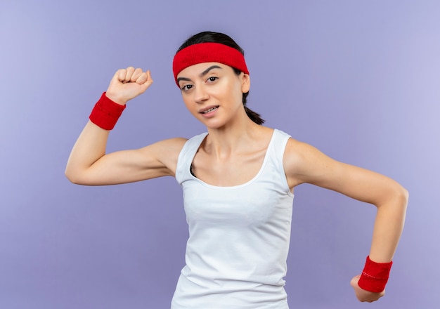 Mujer joven fitness en ropa deportiva con diadema sonriendo alegremente levantando puños feliz y positivo de pie sobre la pared púrpura